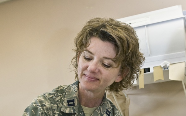 A U.S. Air Force female officer, wearing ABU (Airman Battle Uniform) with captain's rank insignia, is working in what appears to be a medical or occupational therapy setting. She is fitting or adjusting a white orthotic/splint device on a patient's hand or wrist. The setting appears to be a clinical examination room.