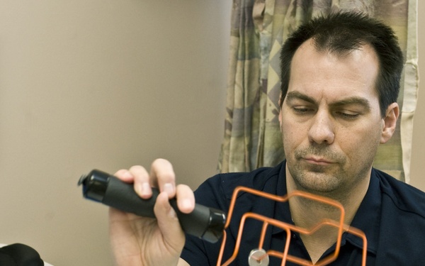 A man in a dark blue polo shirt is examining or treating a foot/ankle using a handheld medical device, likely in a clinical or therapy setting. An orange light-painted graphic overlay appears on his chest, possibly added in post-processing. The setting includes a treatment table with a blue pad and towel, and a calendar on the wall in the background.