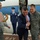 A civilian official in a dark suit and orange tie is being greeted on an airport tarmac, shaking hands warmly with a U.S. Air Force officer in camouflage uniform. The scene appears to be an official arrival ceremony at an Air Force Base, conducted in rainy conditions. Additional military personnel are present, including a uniformed honor guard and another officer holding an umbrella.