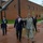 A group of individuals, including a military officer in Air Force camouflage uniform and several men in business suits, are walking together on a brick plaza outside a red-brick institutional building. The military officer appears to be gesturing and directing attention toward something off-frame, suggesting a guided tour or official visit. A large missile or rocket display is mounted on a pedestal in the foreground area.