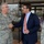 Two individuals are shaking hands in front of what appears to be a military facility entrance. One person is wearing a US Air Force camouflage uniform with the name tape 'SELVA' and general officer rank insignia, while the other is dressed in a civilian navy blue suit with an orange tie. A third uniformed individual with the name tape 'FRASER' is partially visible on the right side of the frame.