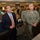 The image shows a group of individuals, including civilians in business attire and military personnel in U.S. Air Force camouflage uniforms, gathered in what appears to be a military operations or command center. The foreground features a civilian man in a navy suit with an orange tie and a U.S. Air Force officer with visible rank insignia and a name tag reading 'SELVA'. The setting includes multiple computer workstations, monitors, and a busy background of additional personnel.
