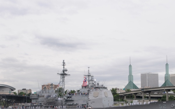 A U.S. Navy warship bearing hull number 52 is navigating a river through an urban area, accompanied by a red tugboat. Sailors in white dress uniforms are lined up along the ship's deck in a formal 'manning the rails' formation. The background features a recognizable cityscape with distinctive green spired structures consistent with the Oregon Convention Center in Portland, Oregon.