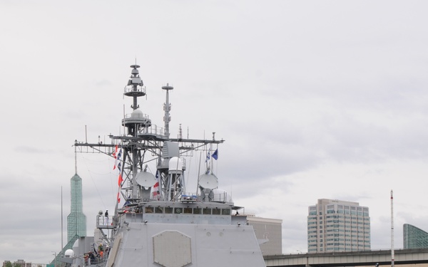 A U.S. Navy guided-missile cruiser bearing hull number 52 is maneuvering in an urban waterway, assisted by a tugboat named 'Vancouver'. Sailors in white dress uniforms are manning the rails in a formal ceremonial formation, suggesting an arrival or departure event. The city skyline in the background, including a distinctive green glass tower, is consistent with Portland, Oregon.