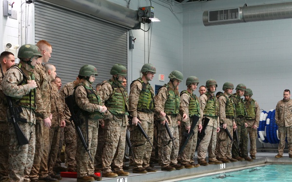 A group of military personnel in full combat gear, including helmets, tactical vests, and rifles, are lined up along the edge of an indoor swimming pool. An instructor in red swim trunks stands in the pool demonstrating or giving instructions, while two additional personnel in standard uniforms observe from the side. This appears to be a military water survival or combat swimming training exercise.