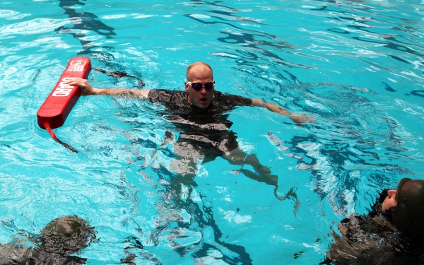 Three individuals are in an indoor swimming pool, appearing to conduct water rescue or lifeguard training. One person in the center is holding up a red rescue buoy/torpedo buoy while wearing swim goggles, extending it toward another person. All individuals appear to be wearing dark clothing, consistent with military or professional training attire.