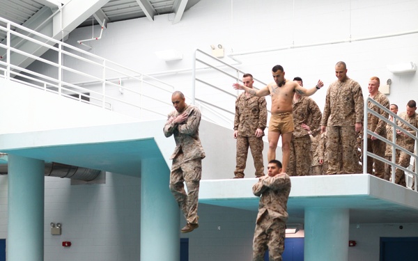 Military personnel in camouflage uniforms are conducting a water survival training exercise at an indoor pool facility. Two individuals are captured mid-jump from an elevated platform approximately 13 feet above the pool, while a group of uniformed personnel wait their turn on the platform. Military gear and backpacks are visible along the pool deck below.