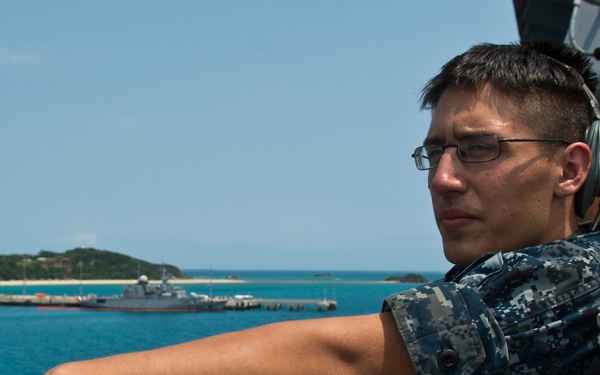 A U.S. Navy sailor wearing Navy Working Uniform (NWU) digital camouflage and a communications headset leans against a ship's railing, gazing toward a harbor. In the background, a naval vessel is visible docked at a pier near a tropical coastline with turquoise water. A second person in what appears to be Marine Corps MARPAT camouflage is partially visible behind the sailor.