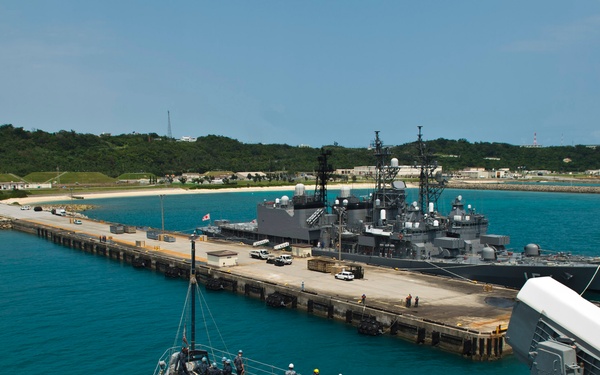 A naval warship is approaching or departing a military pier where another warship is already moored. The scene appears to be at a tropical naval base, identifiable by the turquoise water, sandy beach, and lush green hillside in the background. Crew members are visible on the forecastle deck of the foreground vessel, likely preparing mooring lines.
