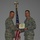 Two U.S. Air Force personnel in Airman Battle Uniform (ABU) camouflage pattern are engaged in a formal award or recognition ceremony. One individual is presenting a certificate or award document to the other, with both holding the document simultaneously in a handshake-style presentation. A U.S. flag on a staff with a gold eagle finial is visible in the background.