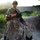 A soldier in MultiCam/MTP camouflage uniform stands against a mud/adobe wall while holding a leash attached to a military working dog (Belgian Malinois or dark German Shepherd) lying on the ground. The soldier is equipped with a helmet, ballistic eyewear, body armor, and an assault rifle. The setting appears to be a rural compound environment consistent with operations in Afghanistan or a similar arid region.