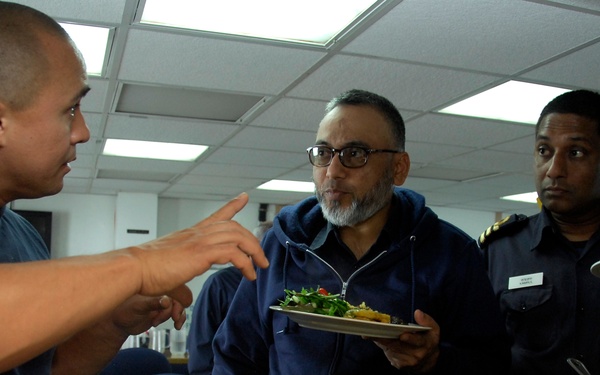 Three men are gathered at what appears to be a food service counter or buffet line, likely in a military or institutional setting. A man on the left is gesturing and speaking while serving food from metal trays, while two men on the right are holding plates with food. The man on the far right appears to be wearing a uniform with rank insignia and a name tag.