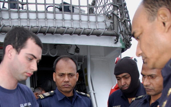 A U.S. Coast Guard member is demonstrating or instructing foreign naval personnel on the use of a fire hose nozzle aboard a vessel. The group appears to be engaged in a joint training or capacity-building exercise on the ship's deck. Multiple uniformed personnel with name tags and rank insignia are attentively observing the equipment demonstration.