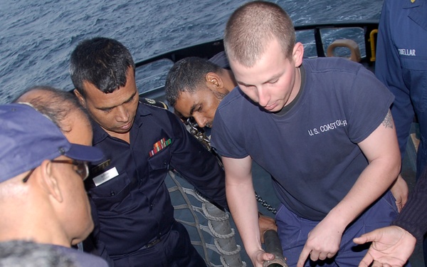A U.S. Coast Guard member in a gray 'U.S. COASTGUARD' t-shirt is demonstrating or instructing a group of foreign naval/maritime personnel on the use of equipment, likely a fire hose nozzle or similar maritime tool. The setting is the deck of a vessel at sea, suggesting a joint training or capacity-building exercise. The foreign personnel are wearing dark navy uniforms with rank insignia and ribbons.