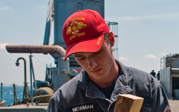 A U.S. Navy sailor identified as 'Newman' is working on the deck of USS Tortuga (LSD-46), handling a wooden board near a pipe or tube. He is wearing dark blue coveralls, yellow work gloves, and a red cap embroidered with 'USS Tortuga LSD-46 Flying Squad.' The setting is the open deck of a naval vessel at sea under clear skies.