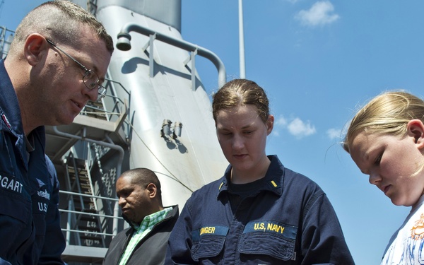 Two U.S. Navy personnel are on the deck of a naval vessel demonstrating rope-tying or knot techniques to civilians, including a young girl. The setting appears to be an open-ship event or educational tour aboard a military vessel. The scene takes place on a sunny day with clear blue skies visible in the background.