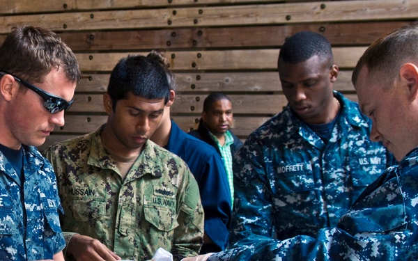 U.S. Navy personnel are going through a food service line, likely at an outdoor or hangar-bay cookout or morale event. Sailors in Navy Working Uniforms (NWU) are being served food including lettuce, burgers or sandwiches, and condiments. Name tapes visible include RIDDLE, HUSSAIN, and MOFFETT.