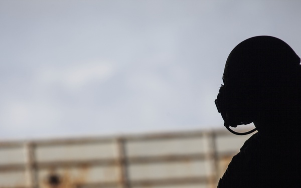 The image shows silhouetted figures against a light sky, with the primary subject wearing a helmet and communications headset/oxygen mask equipment. A large corrugated metal structure or barrier is visible in the background, slightly out of focus. The strong backlighting creates a dramatic silhouette effect, obscuring identifying details.