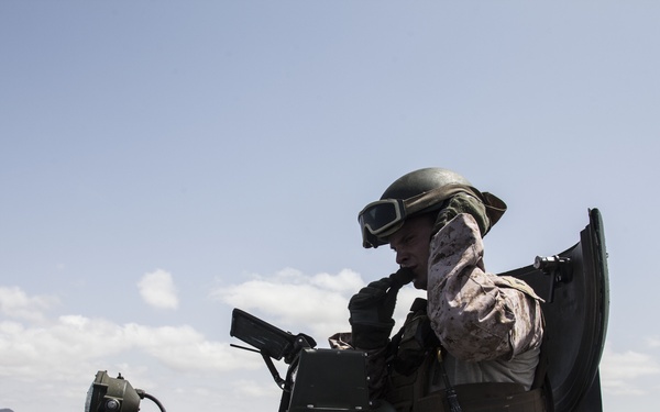 A military personnel is positioned in the open hatch of an amphibious assault vehicle (AAV) traveling on open water. The soldier is wearing desert MARPAT camouflage uniform, a combat helmet with mounted goggles, and appears to be communicating via radio. A coastline with arid terrain is visible in the background.