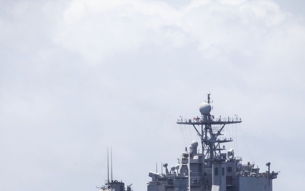 A large gray military amphibious warfare ship is photographed at sea under an overcast sky. The vessel features a prominent flight/vehicle deck extending from the bow, a tall superstructure with radar and communications equipment, and an open well deck or vehicle bay visible at the stern. Personnel are visible standing along the deck railing.