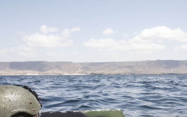 A military personnel wearing a combat helmet with communications headset is positioned inside what appears to be an Amphibious Assault Vehicle (AAV) traveling across open water. The vehicle is moving toward an arid, mountainous coastline visible in the background. The scene suggests an amphibious military operation or training exercise.