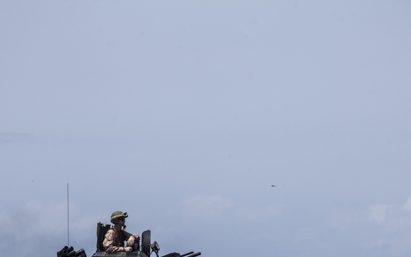 The image shows an amphibious assault vehicle (AAV) traversing open ocean water, with military personnel visible in the turret and on the hull. The vehicle is painted in olive drab/dark green and is creating a wake as it moves through choppy blue water. This appears to be a military amphibious operation or training exercise.