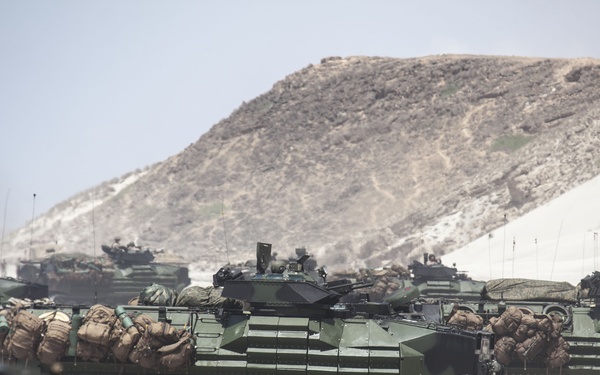 The image shows a formation of multiple Amphibious Assault Vehicles (AAVs), likely AAV-7A1s used by the U.S. Marine Corps, staged on a sandy beach or coastal terrain. The vehicles are loaded with military gear and personnel, suggesting a amphibious landing exercise or operation. A arid, rocky hillside is visible in the background under a hazy sky.