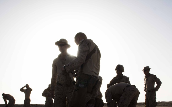 A group of military personnel are gathered in an arid, desert environment, silhouetted against a bright sky suggesting strong backlighting from the sun. The soldiers appear to be conducting equipment checks, gear preparation, or a field inspection. The scene suggests a training exercise or pre-mission staging activity.