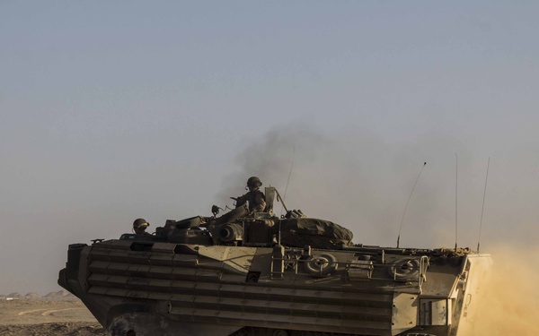 An amphibious assault vehicle (AAV) is moving at speed across an arid desert terrain, kicking up a large dust cloud from its tracks. At least two crew members are visible in open hatches on top of the vehicle. The scene appears to be a military training exercise or operational movement in a desert environment.