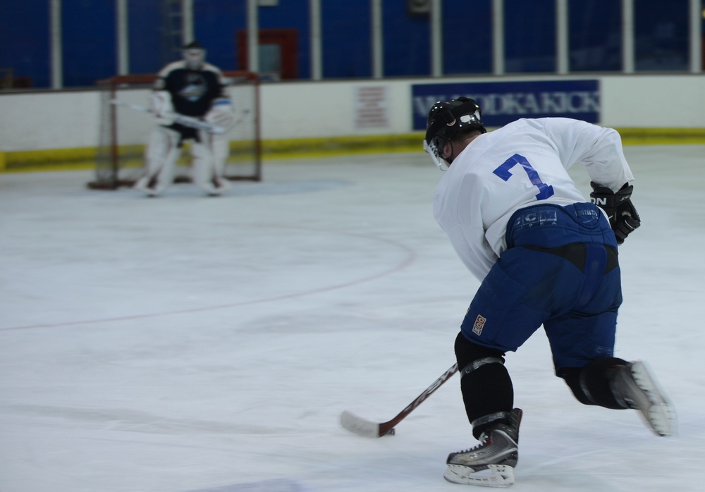 An ice hockey player wearing number 7 in a white jersey and blue pants is captured mid-shot on an indoor ice rink, skating toward the goal. A goaltender in a dark jersey is positioned in the net in the background, ready to defend. The image was taken at an indoor hockey arena with blue and white arena boards visible.