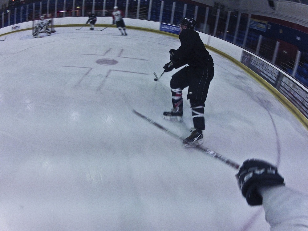 A GoPro-style first-person perspective shot taken during an ice hockey game or practice session inside an indoor rink. The foreground shows the camera operator's gloved hand and stick, while a player in black gear is visible mid-stride in the middle distance. Additional players and a goaltender are visible in the background near the net.