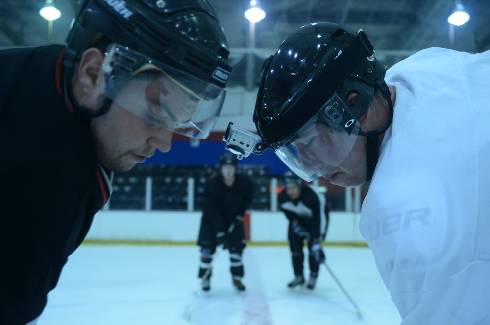 Two ice hockey players in the foreground are facing off or in close confrontation, with their helmeted heads nearly touching. Both wear black helmets with visors and are on an indoor ice rink. Two additional players are visible in the background on the ice.