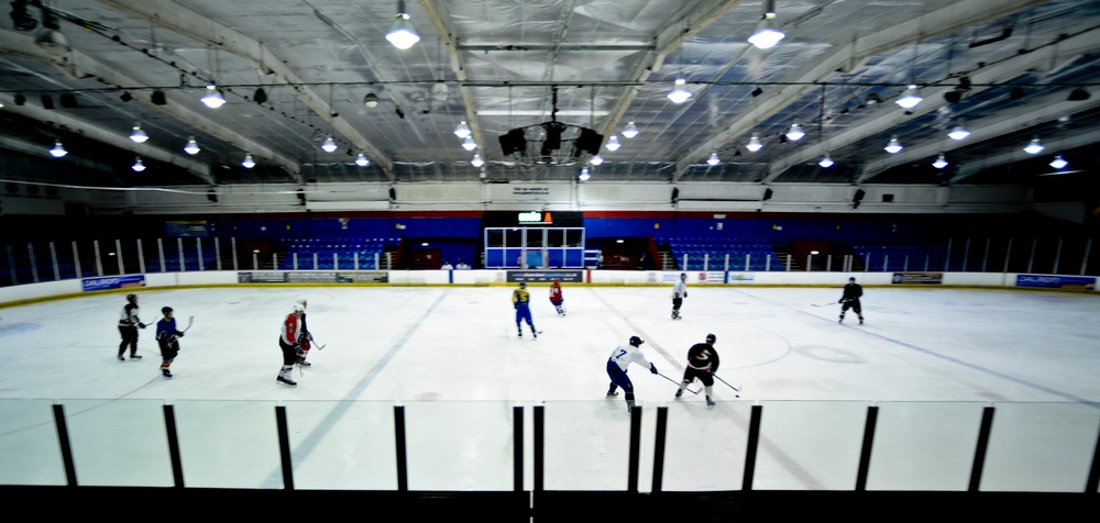 An indoor ice hockey rink captured from an elevated angle behind the glass boards, showing a practice or informal scrimmage session in progress. Multiple players in various colored jerseys are spread across the ice surface, engaged in drills or gameplay. The arena features blue spectator seating in the background, overhead lighting, and standard hockey rink markings.