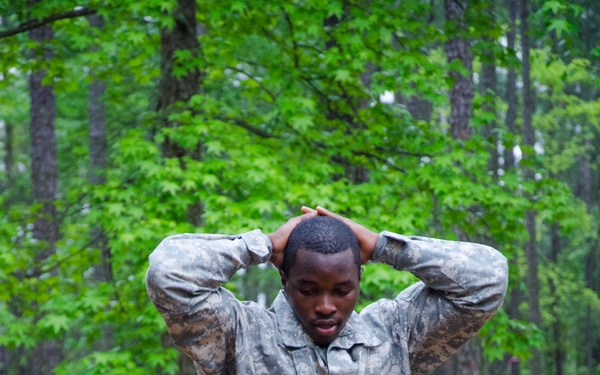 A soldier in US Army ACU (Army Combat Uniform) digital camouflage is navigating a wooden obstacle course, with hands clasped behind his head. The setting appears to be a military training facility in a wooded area, with a second soldier visible in the background. The ground is covered with wood chips, typical of obstacle course installations.