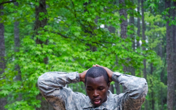 A soldier in US Army ACU (Army Combat Uniform) digital camouflage is navigating a wooden obstacle course in a forested training area. The soldier has both hands clasped behind their head, suggesting a specific movement drill or low-crawl obstacle requirement. Additional soldiers in uniform are visible in the background among the trees.