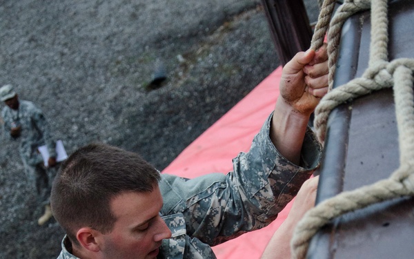 A soldier in US Army ACU (Army Combat Uniform) digital camouflage is climbing a rope ladder attached to a vertical wall structure, viewed from above. Below, orange safety mats are positioned on the ground as fall protection. Additional personnel in uniform are visible in the background observing or supervising.