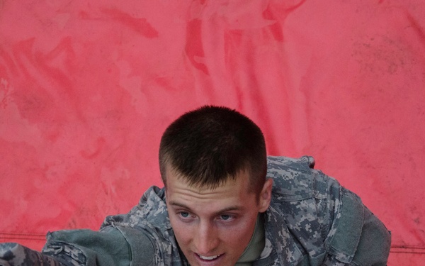A young male soldier in US Army ACU (Army Combat Uniform) digital camouflage is climbing over a rope obstacle, likely during a military training exercise or obstacle course. His expression shows physical exertion and determination as he reaches forward with one hand while gripping a thick rope with the other. A red tarp or barrier forms the background behind him.
