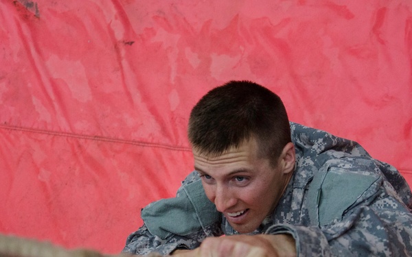 A young male soldier in US Army digital camouflage (ACU pattern) is crawling or pulling himself along a rope obstacle course element. He appears to be smiling or grimacing with effort, suggesting this is a training exercise or competition. A red tarp or mat is visible in the background and beneath him.