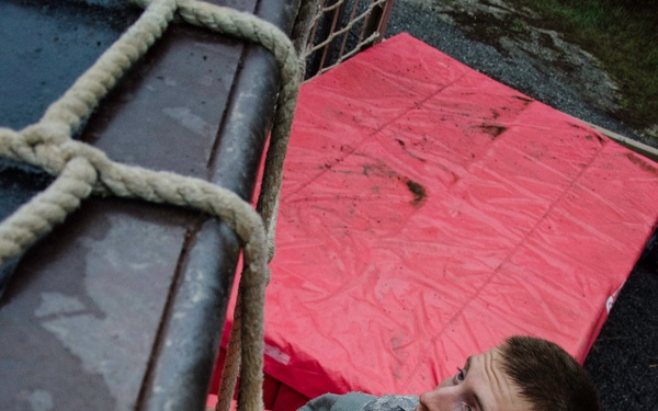 A soldier in US Army ACU (Army Combat Uniform) digital camouflage is climbing a rope ladder or cargo net on what appears to be a military obstacle course. The photo is taken from above, looking down at the climber who is gripping the ropes tightly and looking upward. Red safety crash mats are visible on the ground below.