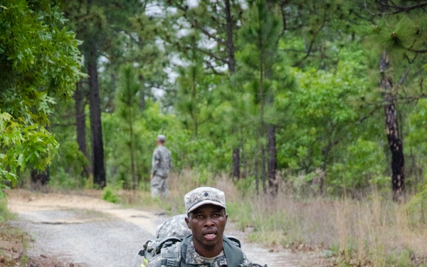 A U.S. Army soldier in ACU (Army Combat Uniform) is running along a gravel path through a wooded training area while carrying full gear. The soldier is equipped with a rucksack, tactical vest, and what appears to be an M16/M4-style rifle with a yellow training muzzle cap. A second soldier in uniform is visible in the background, standing on the same path.