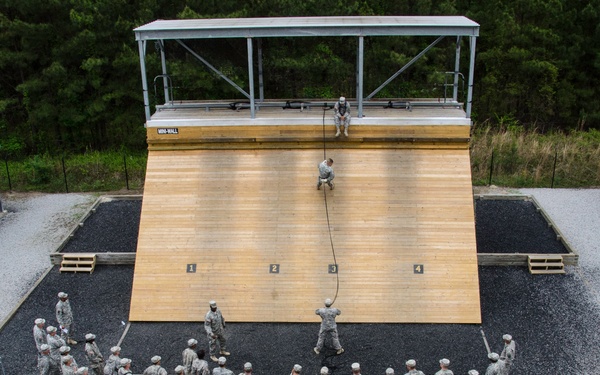 An aerial view of a military training facility showing soldiers gathered around a large rappelling wall. One soldier is actively rappelling down the wooden wall face while an instructor or safety observer sits at the top platform. A large group of uniformed soldiers in ACU (Army Combat Uniform) pattern are assembled in a semicircle watching the demonstration.