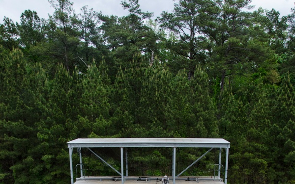 An aerial view of a military rappelling training exercise at what appears to be a purpose-built wooden rappel wall labeled 'MRI-WALL'. One soldier is actively rappelling down the inclined wooden surface while another sits at the top platform, and a third acts as a belayer at the base. A large group of uniformed soldiers in ACU (Army Combat Uniform) pattern stands in formation observing the demonstration.