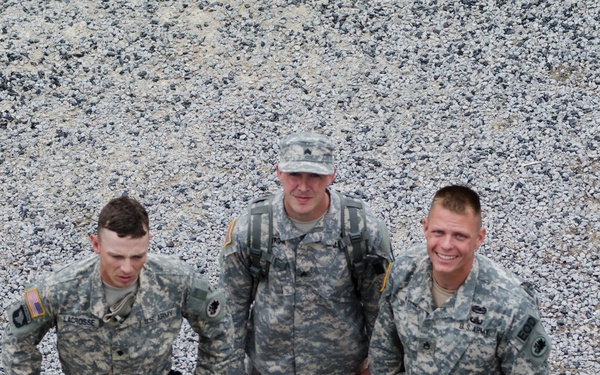 Three U.S. Army soldiers in Army Combat Uniform (ACU) pattern are photographed from an elevated angle, standing on a gravel surface near a wooden beam or barrier. The soldiers are equipped with rappelling/climbing gear including carabiners and harnesses. The leftmost soldier's name tape reads 'LACROSSE' and all three wear U.S. Army insignia with shoulder patches.