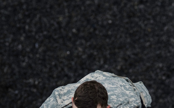 A U.S. Army soldier in Army Combat Uniform (ACU) digital camouflage pattern is crawling or climbing over a low barrier or surface marked with yellow paint lines. The soldier is wearing a U.S. flag patch and an additional unit patch on the left sleeve, suggesting active duty service. The activity appears to be part of a physical training exercise or obstacle course.