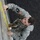 Best Warrior Competition A U.S. Army soldier in ACU (Army Combat Uniform) is performing what appears to be a physical fitness exercise, specifically a wall climb or obstacle course activity. The image is rotated 90 degrees, showing the soldier gripping a wall or barrier with yellow markings while wearing combat boots. The soldier's uniform displays a U.S. flag patch and a unit patch on the left shoulder.