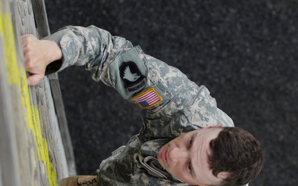 A U.S. Army soldier in ACU (Army Combat Uniform) is performing what appears to be a physical fitness exercise, specifically a wall climb or obstacle course activity. The image is rotated 90 degrees, showing the soldier gripping a wall or barrier with yellow markings while wearing combat boots. The soldier's uniform displays a U.S. flag patch and a unit patch on the left shoulder.