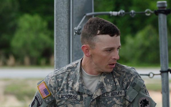 Two U.S. Army soldiers in ACU (Army Combat Uniform) digital camouflage are visible at what appears to be a training facility or obstacle course. The soldier in the background is identified by his name tape as 'LACROSSE' and wears a U.S. Army tape, an American flag patch, and what appears to be an Airborne or similar unit patch on his left shoulder. Both soldiers appear to be equipped with rappelling or climbing harness gear.