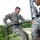 Region 3 Best Warrior Competition A U.S. Army soldier named LACROSSE is preparing to rappel from an elevated training tower, wearing Army Combat Uniform (ACU) with a rappelling harness and carabiner. Several other soldiers in ACUs are visible on the platform observing or assisting. The ground below shows a training area with a tent, tripod-mounted camera, and safety personnel.
