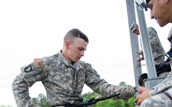 A U.S. Army soldier named LACROSSE is preparing to rappel from an elevated training tower, wearing Army Combat Uniform (ACU) with a rappelling harness and carabiner. Several other soldiers in ACUs are visible on the platform observing or assisting. The ground below shows a training area with a tent, tripod-mounted camera, and safety personnel.