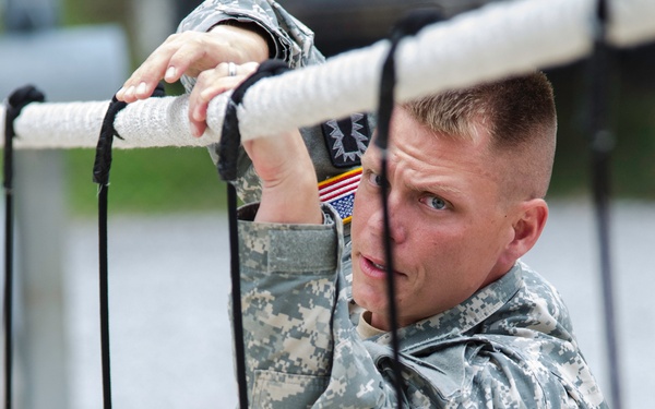 A U.S. Army soldier in Army Combat Uniform (ACU) digital camouflage pattern is gripping a rope or bar obstacle, appearing to be engaged in a physical training or obstacle course activity. The soldier displays a U.S. flag patch and what appears to be a unit patch on the left sleeve. The image is taken at close range with black ropes or cables visible in the foreground.